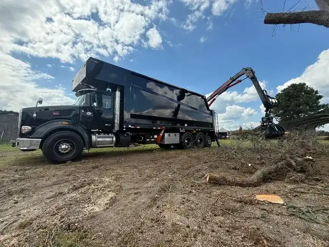Peterbilt grapple truck loading tree debris at a clearing site under blue sky